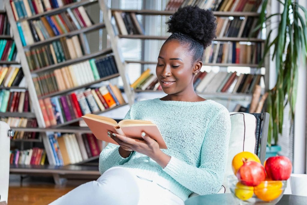 Woman sitting in a library, reading a book and looking at her watch.