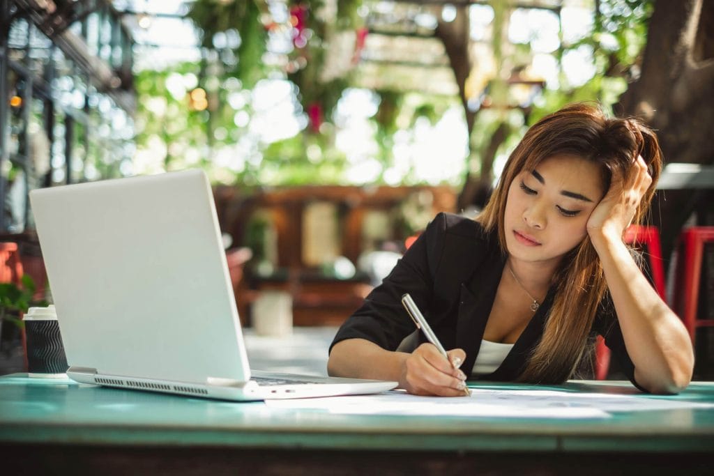 Woman working on a laptop with a notebook beside her on a bright desk.