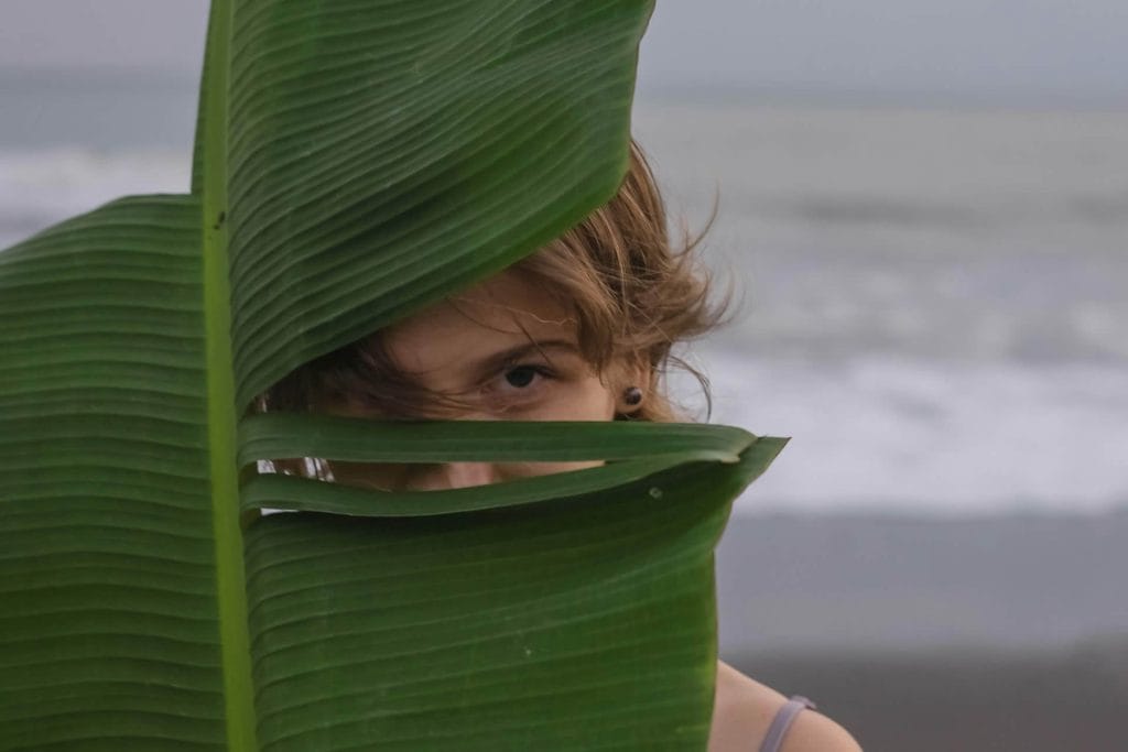 Person peeking playfully through large green leaves.