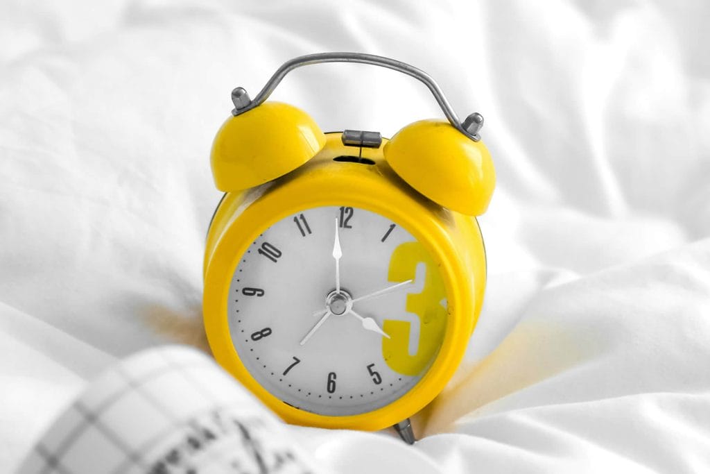 Yellow alarm clock resting on white sheets in soft morning light.